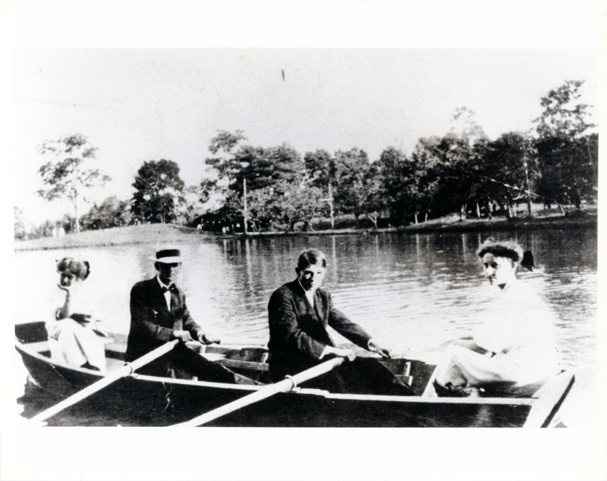 A black and white historic photo showing two men and two women dressed in early 20th century clothing rowing a canoe on a lake
